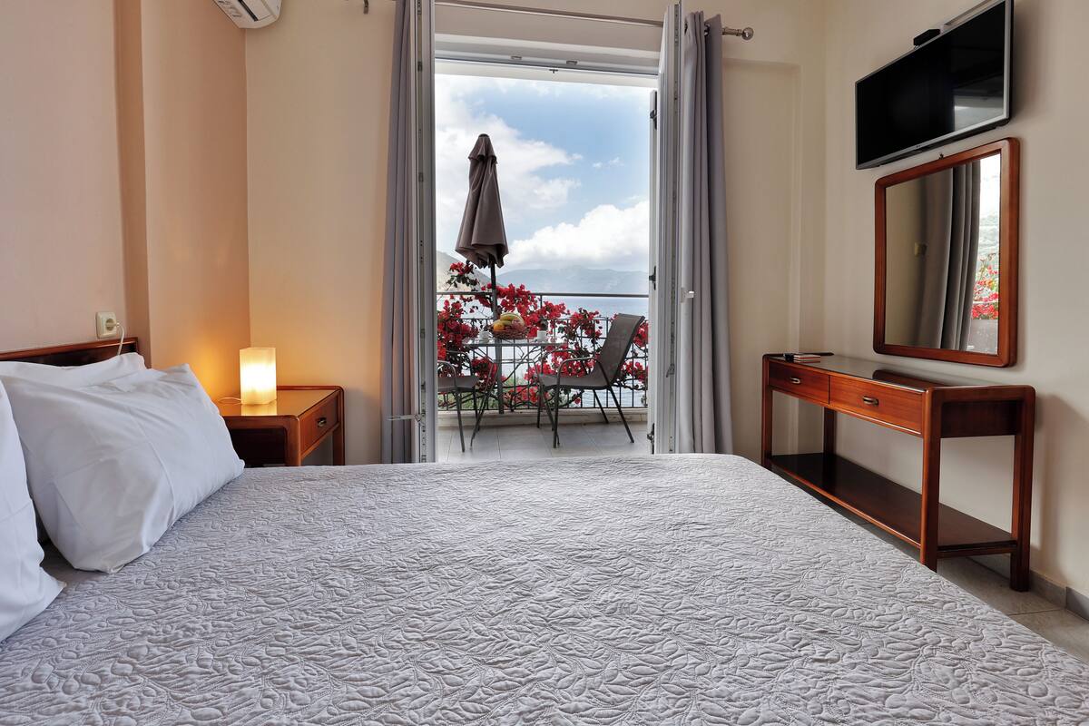 A hotel room with a bed, a bedside table with a lamp, a wooden dresser, a TV, and a balcony with flowers and a chair.