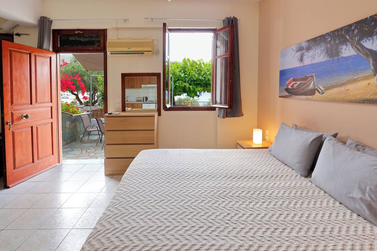 A bedroom with a large bed, a window with a view of greenery, a painting on the wall, and a wooden door.