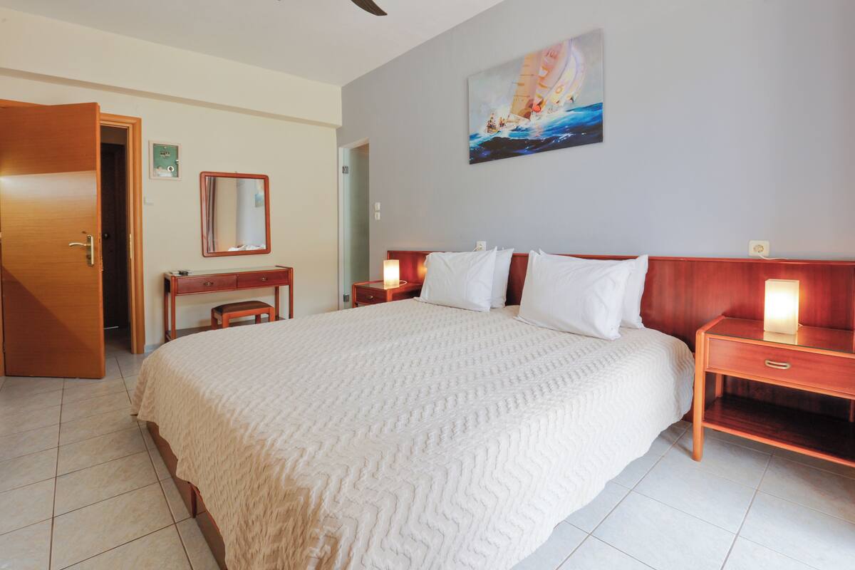 A hotel room with a large bed, bedside tables, a desk, a chair, a mirror, and a painting on the wall.