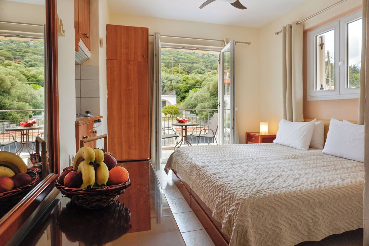 A hotel room with a bed, a dining area with a table and chairs, a fruit basket, and a view of the outdoors.