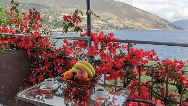 Una mesa con una cesta de fruta, una taza de café y un vaso de agua, con vistas a un paisaje que incluye una masa de agua y colinas.
