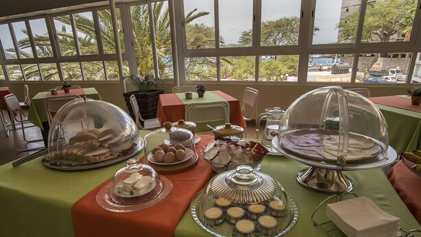 Una mesa de buffet con diversos alimentos cubiertos por campanas de cristal, con vistas a grandes ventanales que muestran palmeras y edificios.