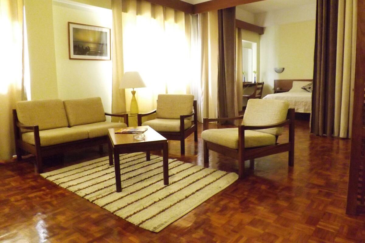 A hotel room with a sofa, armchairs, a small table, a lamp, and a bed.