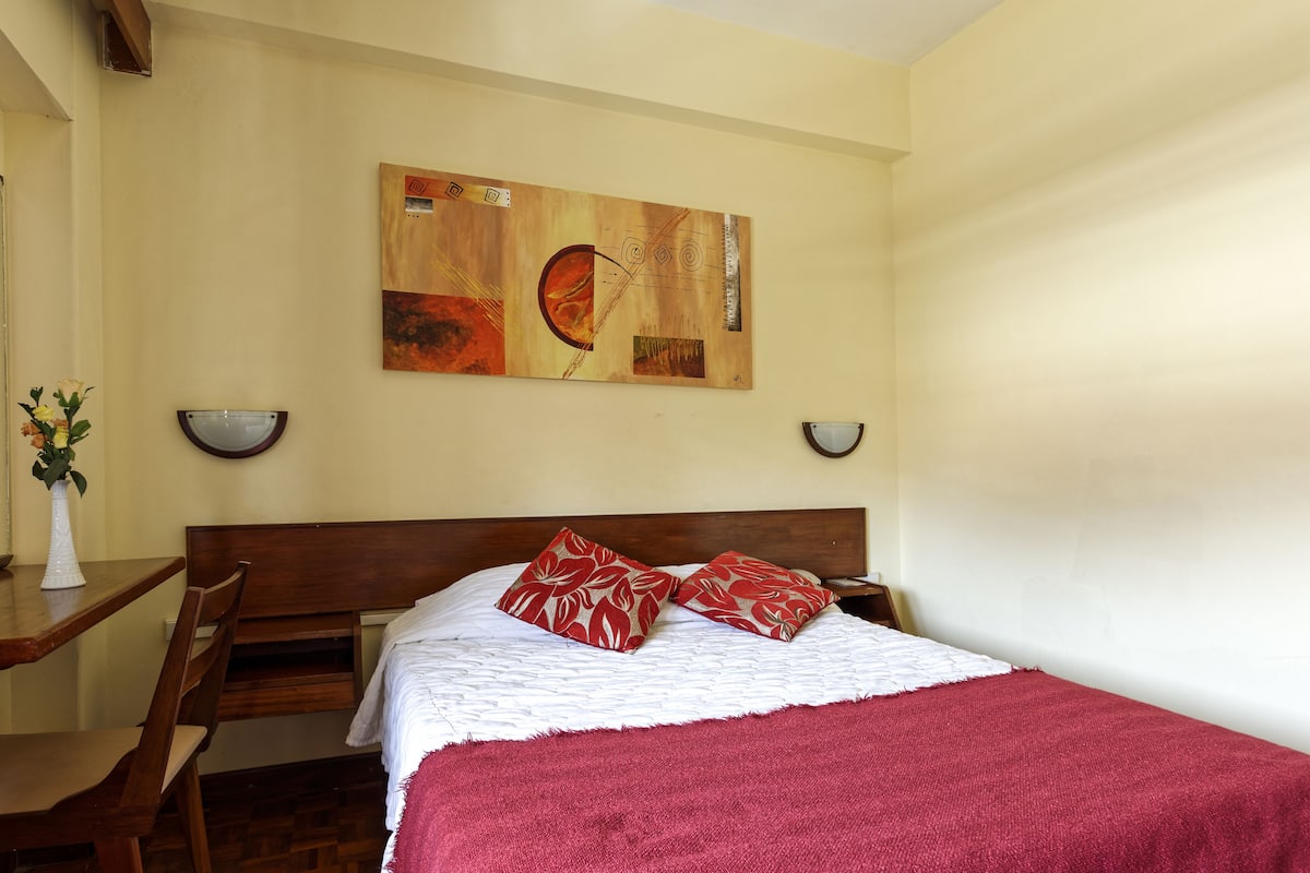 A bedroom with a wooden headboard, a bed with a white and red blanket, two red pillows, a wall-mounted abstract painting, and a small table with a vase of flowers.