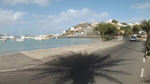 Puerto deportivo con barcos, un paseo marítimo pavimentado y edificios al fondo.