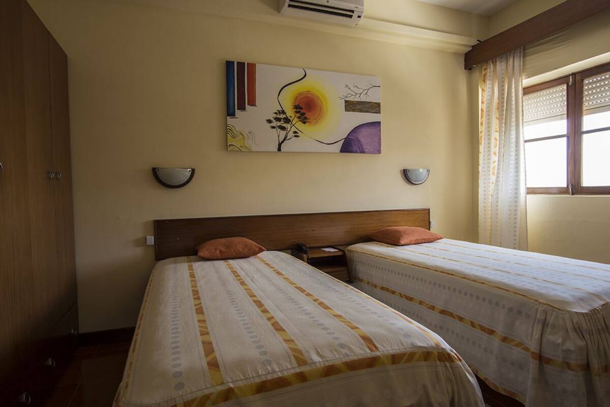 A hotel room with two single beds, a large painting on the wall, and a window with curtains.