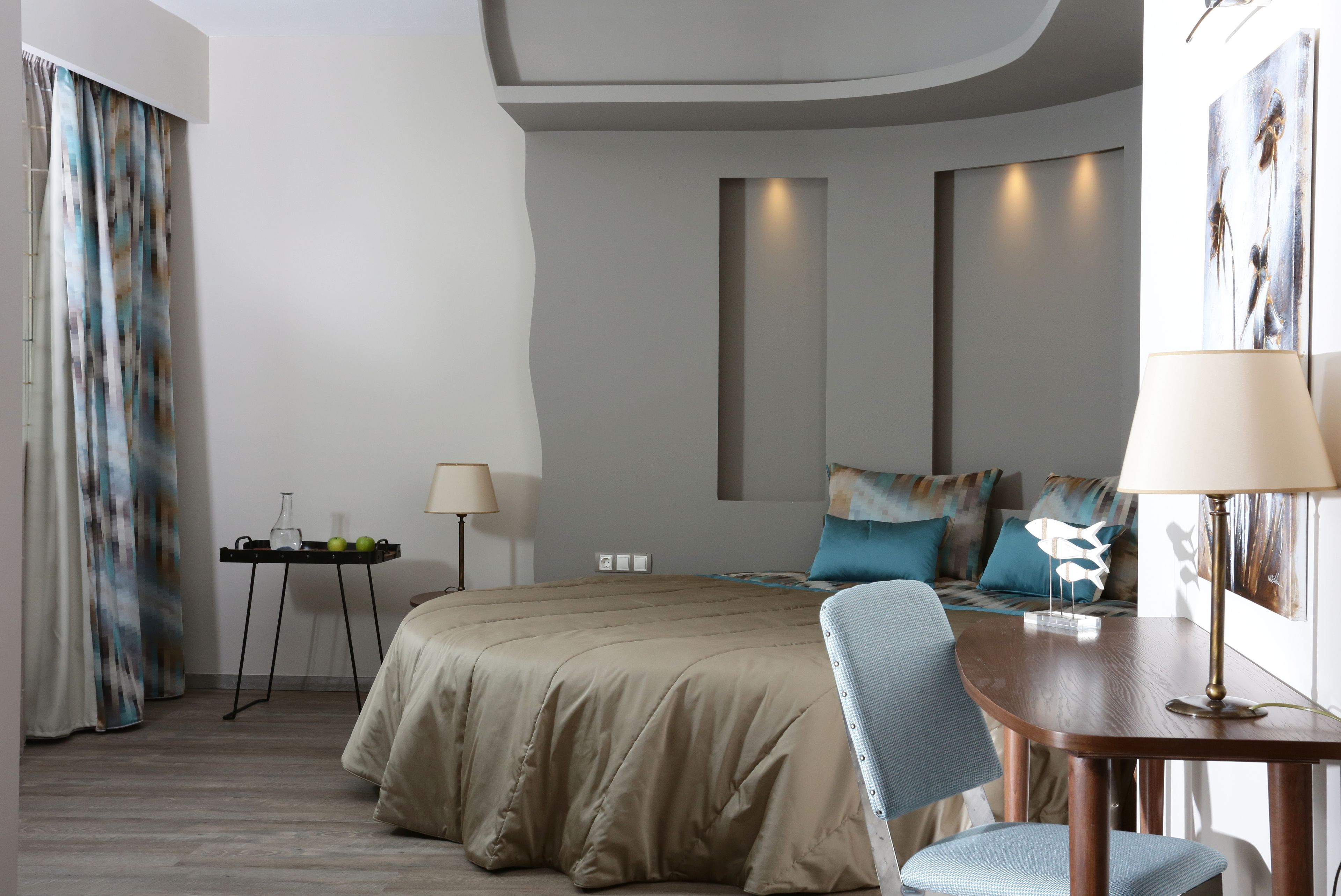 A modern hotel room with a large bed, a desk with a lamp, a small table with a lamp, and a painting on the wall.
