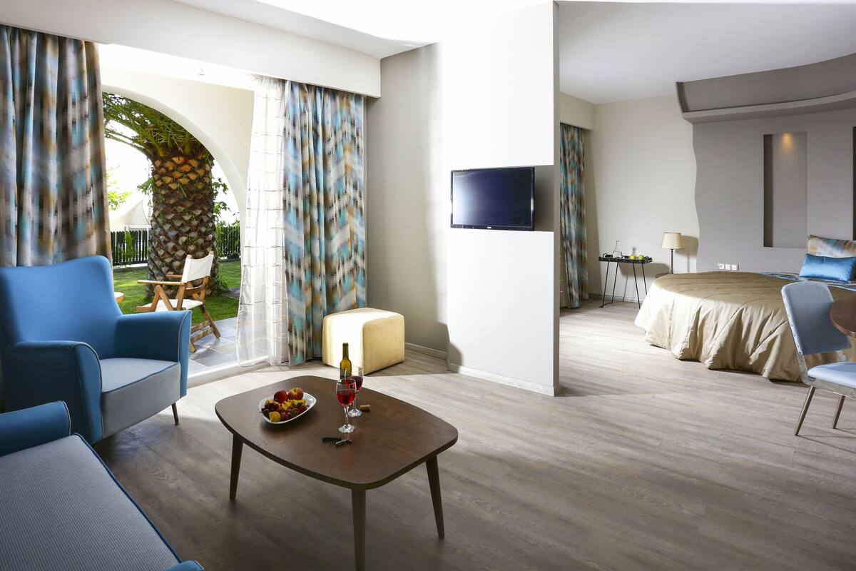 A modern hotel room with a curved ceiling, a flat-screen TV, a bed with blue bedding, a dining area with a table and chairs, and a balcony with a view of a garden and palm tree.