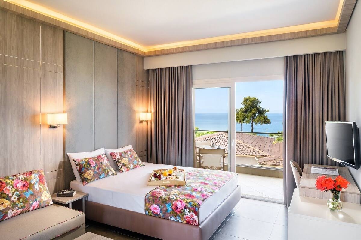 A modern hotel room with a large bed, a TV, and a balcony with a view of the sea.