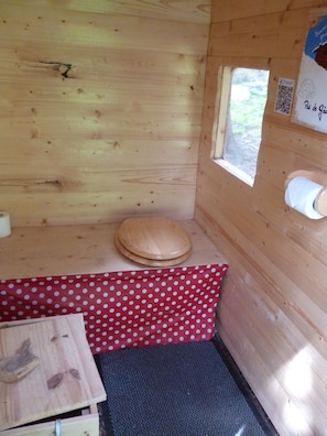 L’intérieur d’une cabane en bois, avec des toilettes en bois, une petite table recouverte d’une nappe à pois rouges et une fenêtre sur laquelle est apposé un code QR.
