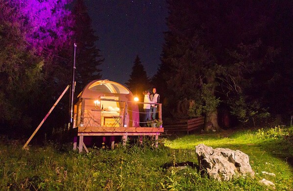 A small, dome-shaped cabin with a wooden deck, surrounded by trees and a rock.