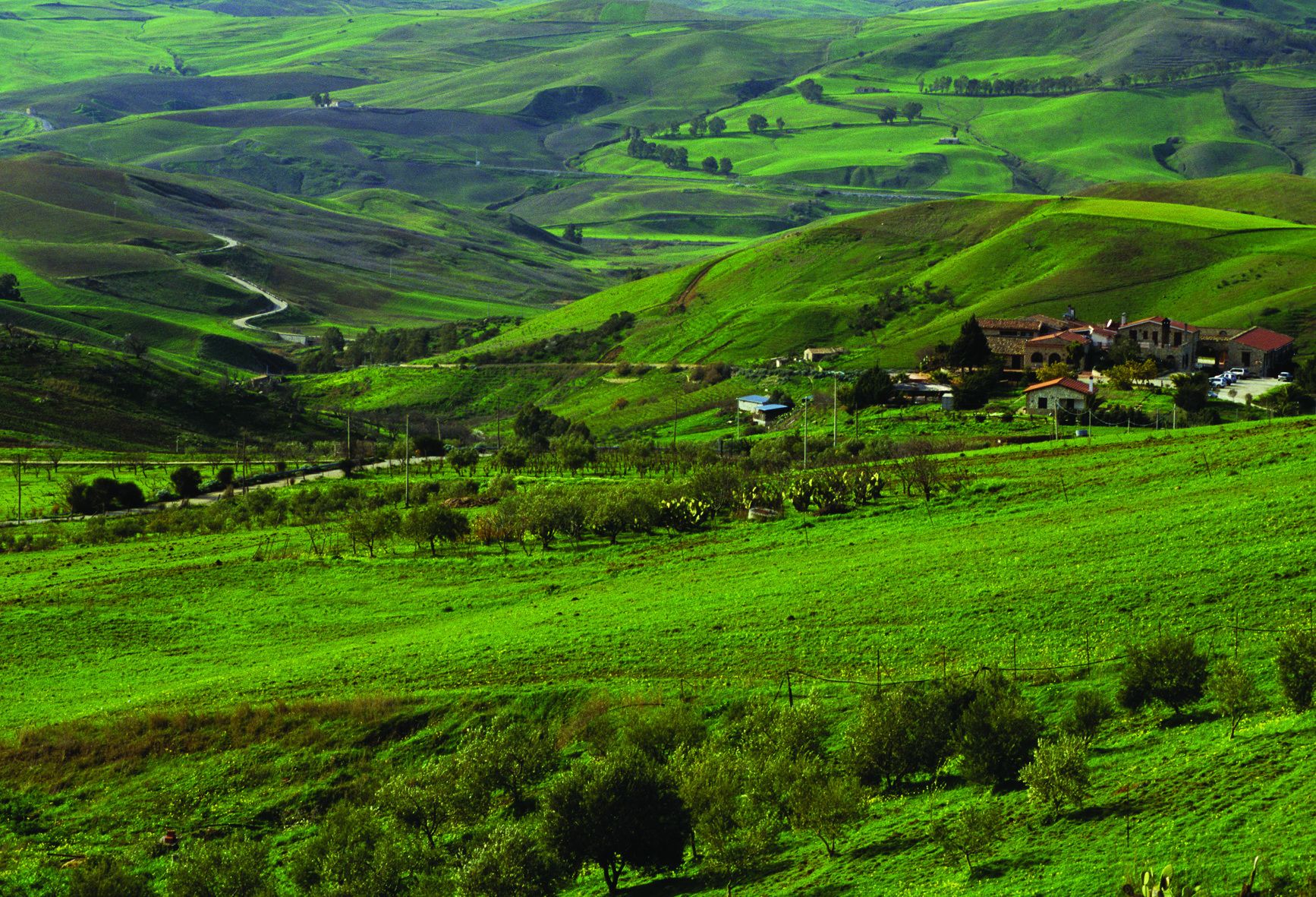 A rural landscape with rolling hills, a winding road, and a cluster of buildings with red roofs.