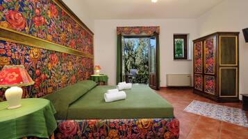 Una stanza con una testiera a motivi floreali, una coperta verde, un comodino con una lampada e vista sul verde attraverso una finestra.