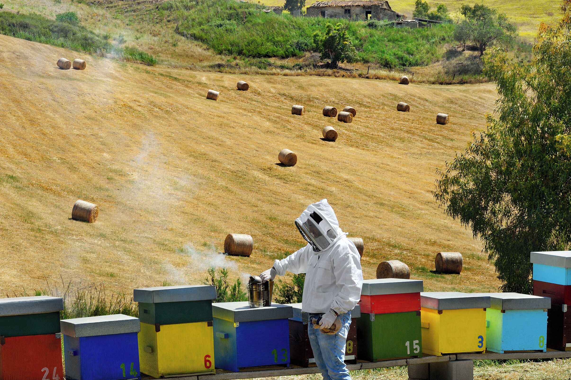 A person in protective gear working near colorful storage units in a rural setting with hay bales in the background.