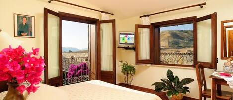 A bedroom with a bed, a TV, a window with a view of a stone wall and hills, and a potted plant.