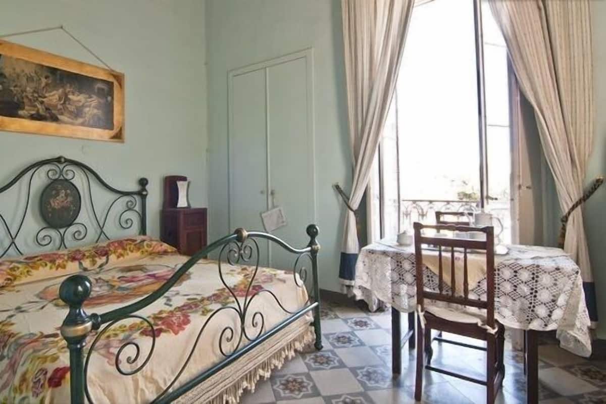 A bedroom with a bed, a chair, a table, a painting, and a window with curtains.