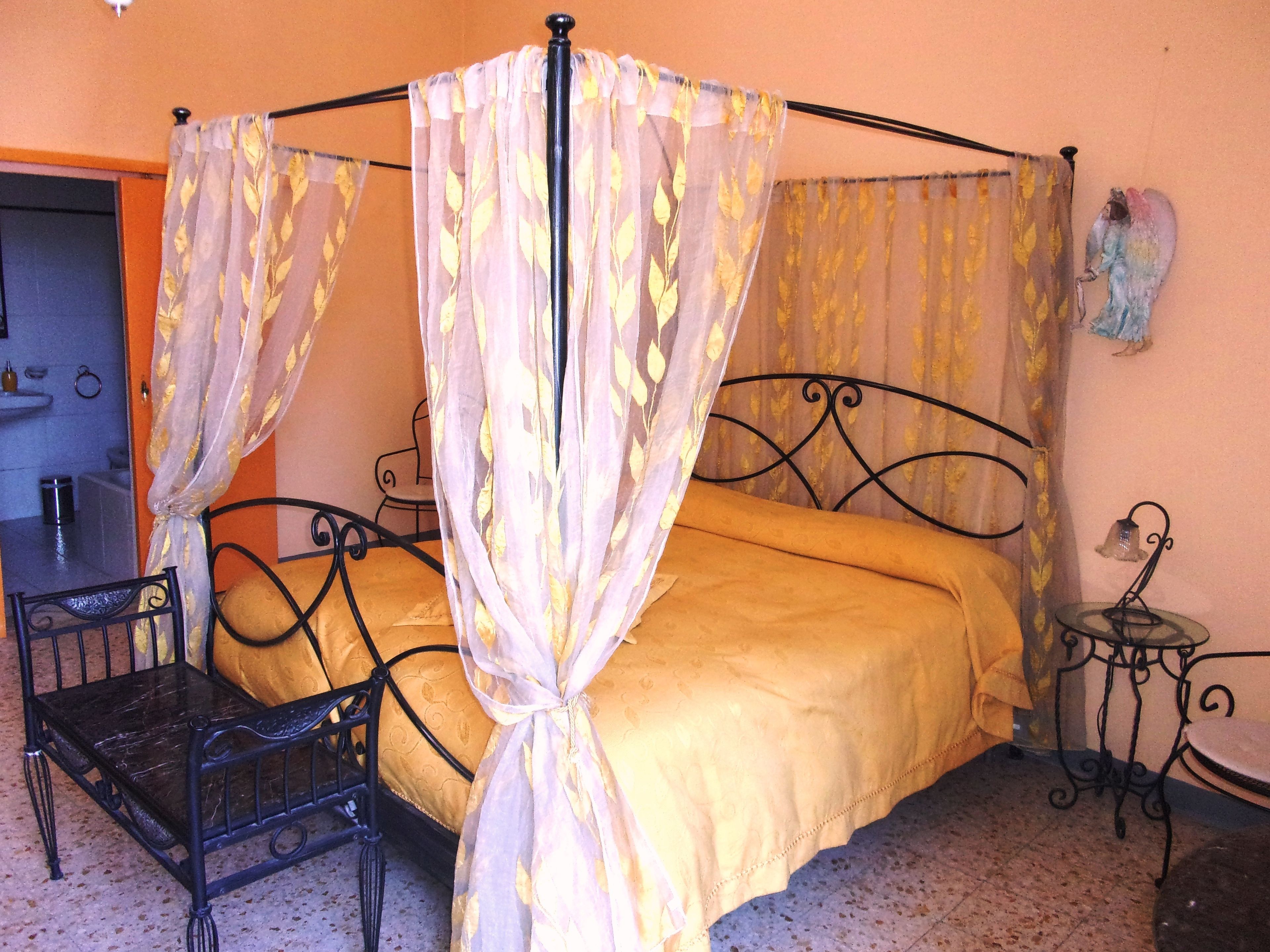 Una cama con dosel, una mesita pequeña, una silla y un estante en la pared.