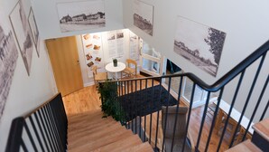 A staircase with wooden steps and a black railing, leading to a room with a white table and chairs, surrounded by framed photographs on the walls.