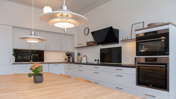 A modern kitchen with white cabinets, a wooden dining table, pendant lights, a built-in oven, and a clock on the wall.