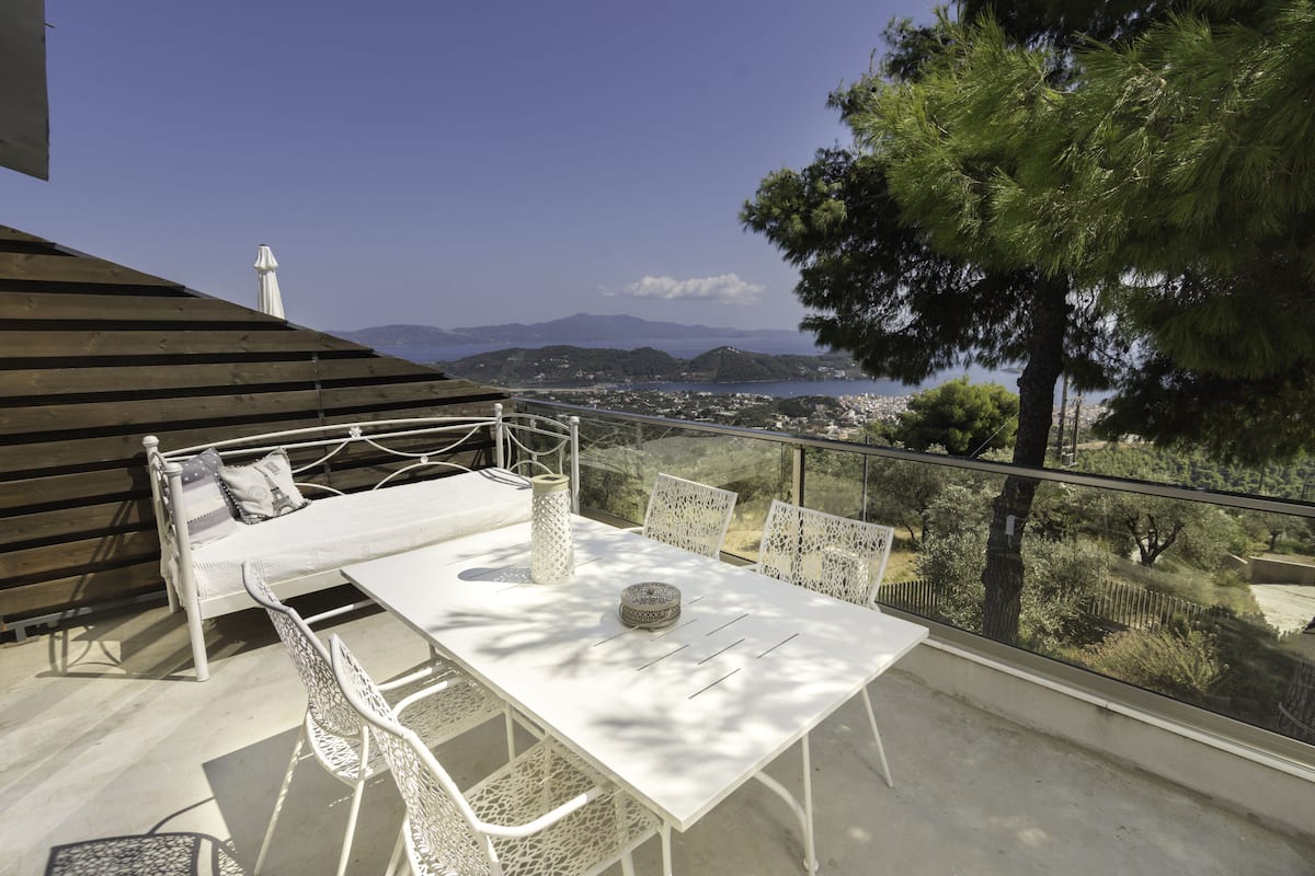 A balcony with a white table and chairs, a bed with pillows, and a view of a coastal town and mountains.