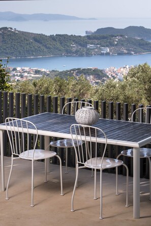 Eine Terrasse im Freien mit Tisch und Stühlen, mit Blick auf eine Küstenlandschaft mit Hügeln und einem Gewässer.