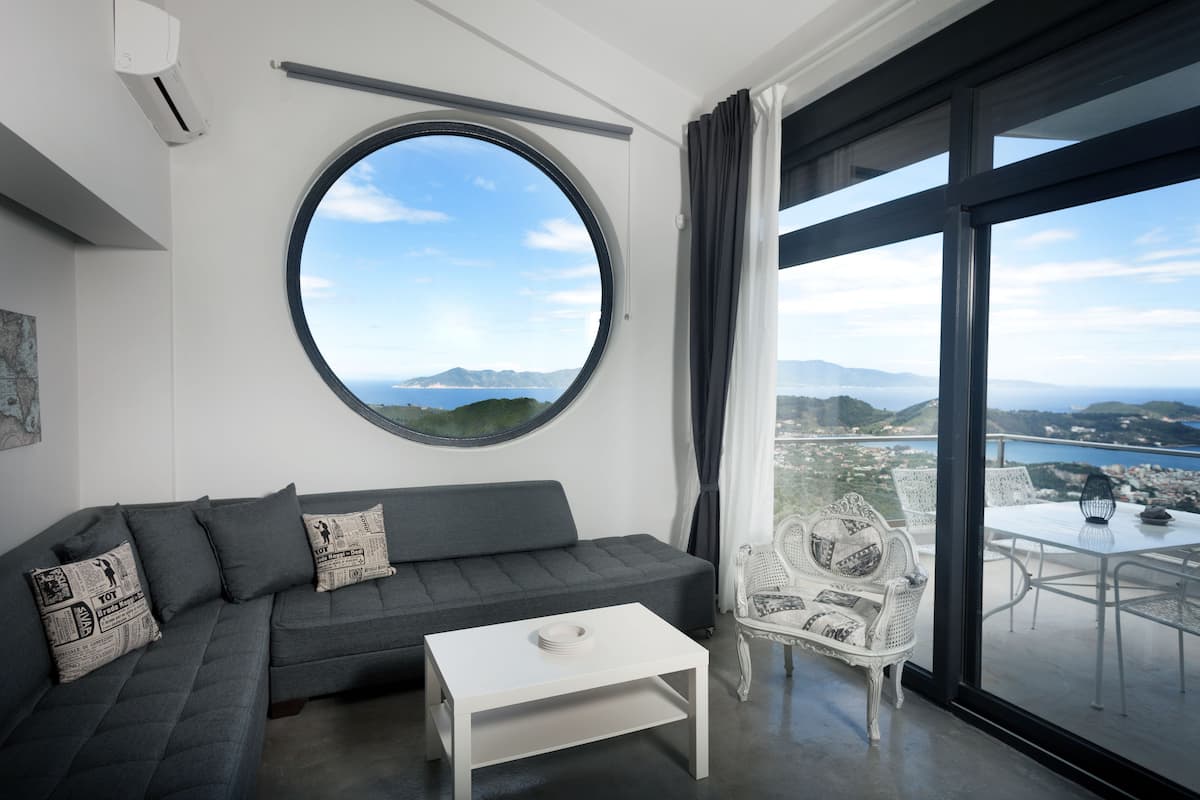 A modern living room with a grey sofa, a white coffee table, and a round window offering a scenic view of the sea and mountains.