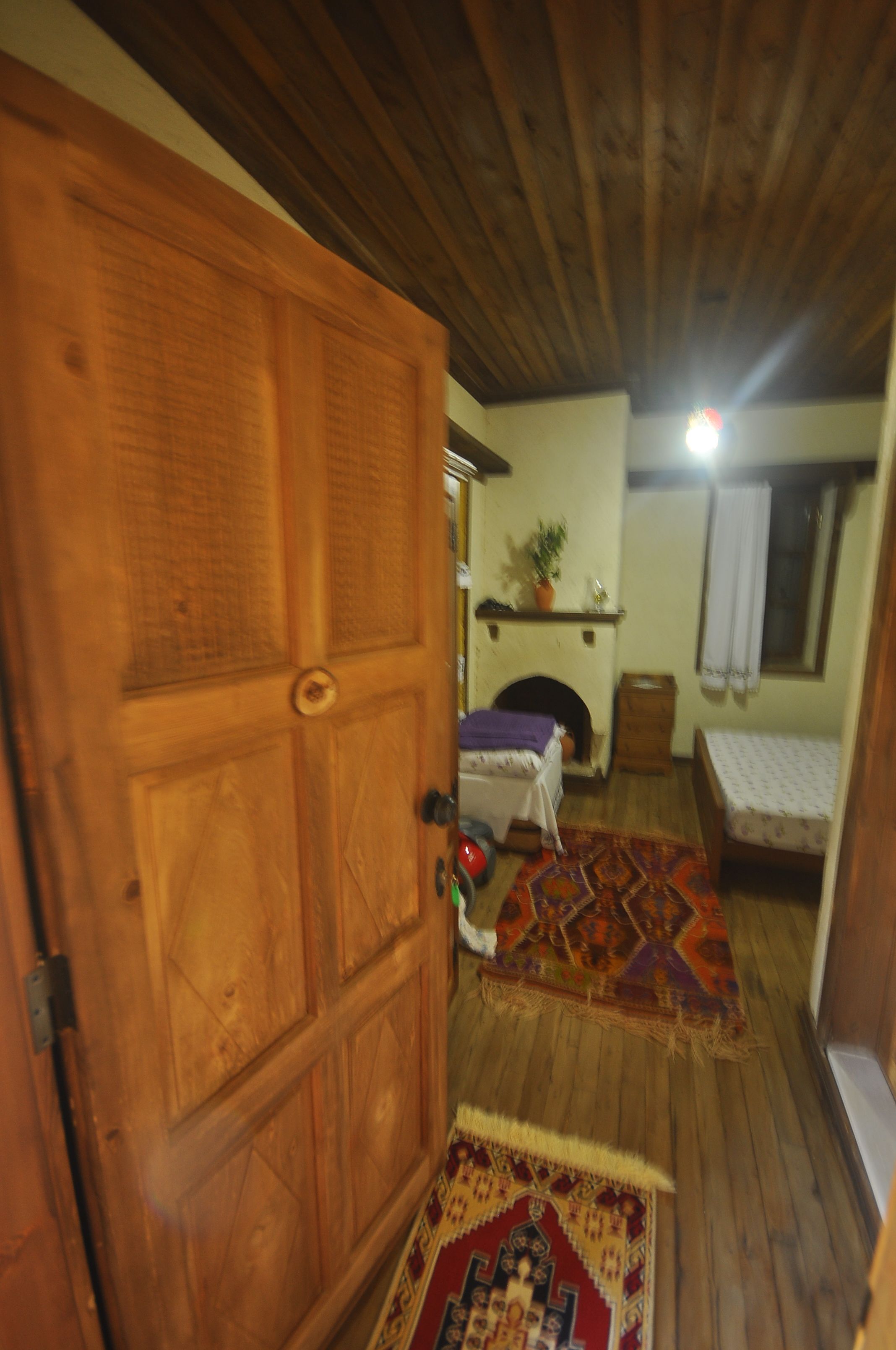 Ein schmaler Flur mit einer Holztür, einem kleinen Tisch mit einer violetten Decke und einem Bett mit einem gemusterten Überwurf, sowie einem Fenster mit weißem Vorhang.