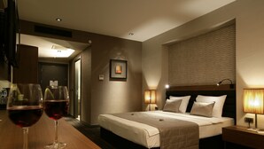 Habitación de hotel con cama, lámparas de noche, un cuadro en la pared y dos copas de vino tinto sobre la mesita.