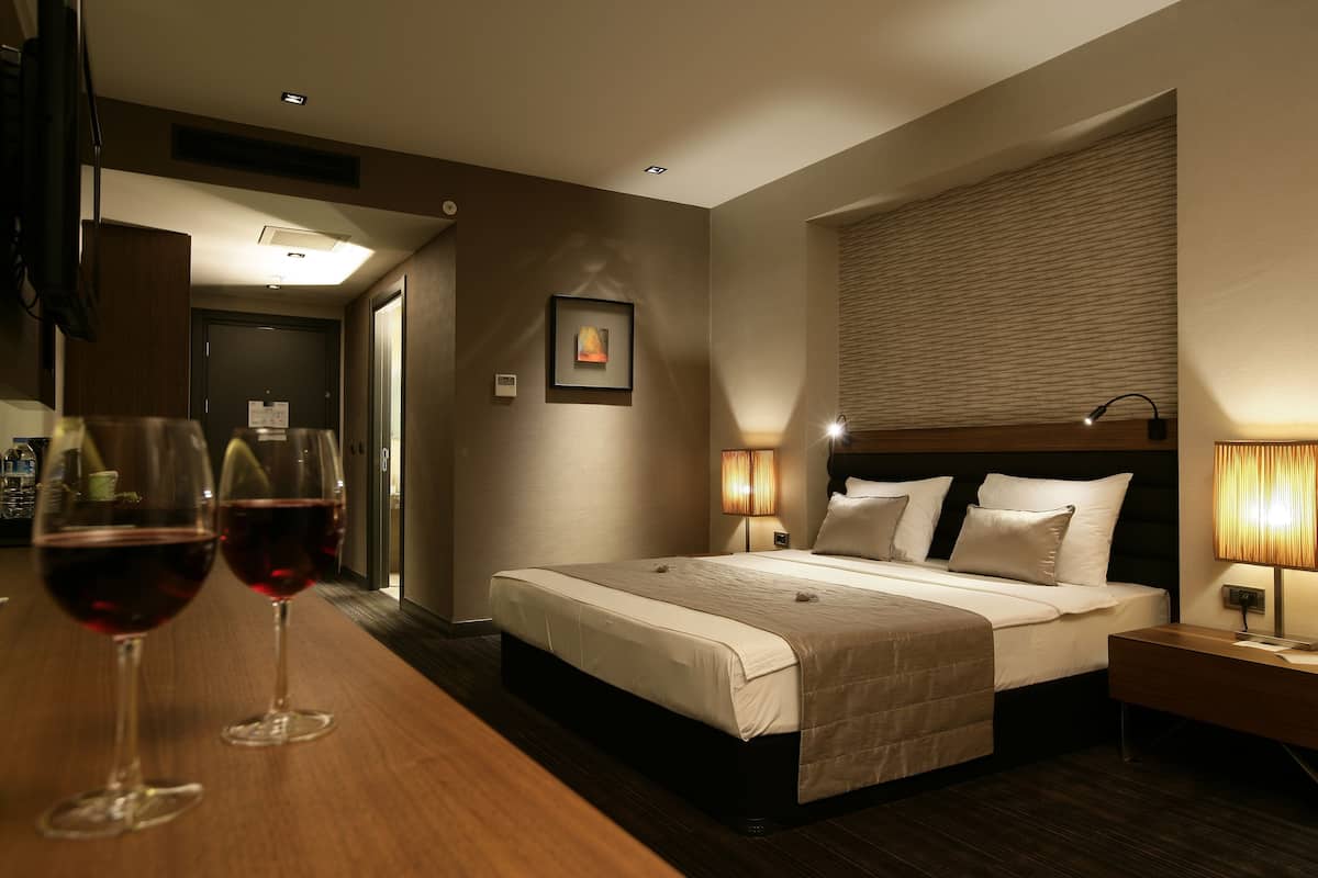 A hotel room with a bed, bedside lamps, a picture on the wall, and two glasses of red wine on the nightstand.