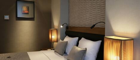 Una cama bien hecha con sábanas blancas y dos almohadas, una lámpara de pared y una obra enmarcada en la pared.