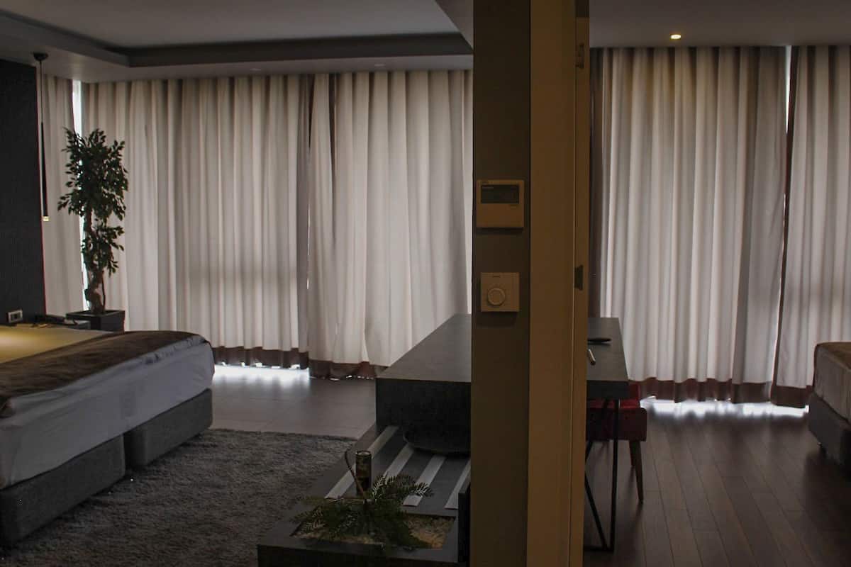 A modern hotel room with a large bed, a desk, and a view of the city through floor-to-ceiling windows.