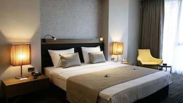 Una habitación de hotel moderna con una cama grande, dos mesitas de noche con lámparas, una silla y vistas al exterior.