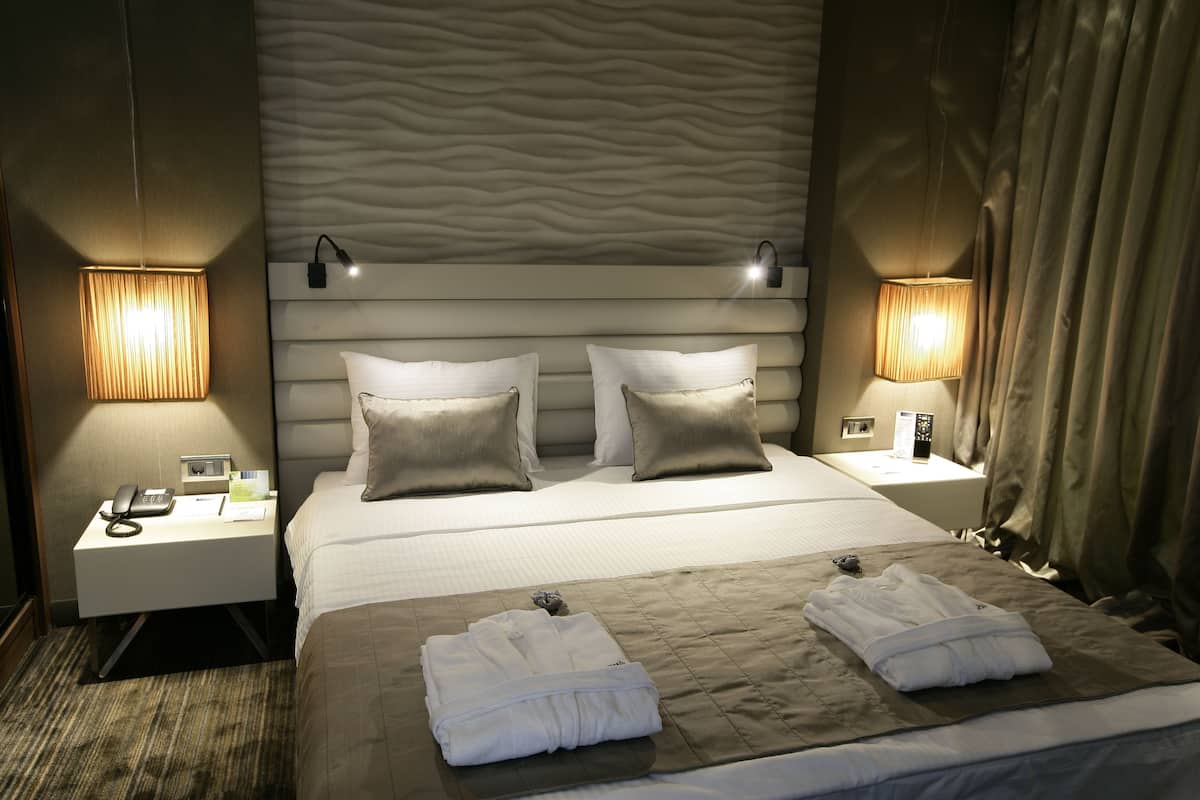 A hotel room with a neatly made bed, two bedside lamps, a nightstand with a phone and a lamp, and a textured wall behind the bed.