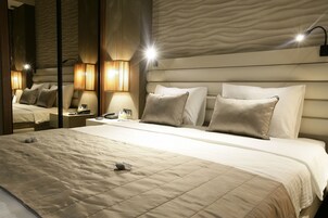 Una cama bien hecha con cubrecama acolchada, una mesita de noche con lámpara y un espejo que refleja el interior de la habitación.