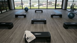 Un gimnasio con equipo de ejercicio, ventanas amplias y vistas a edificios.