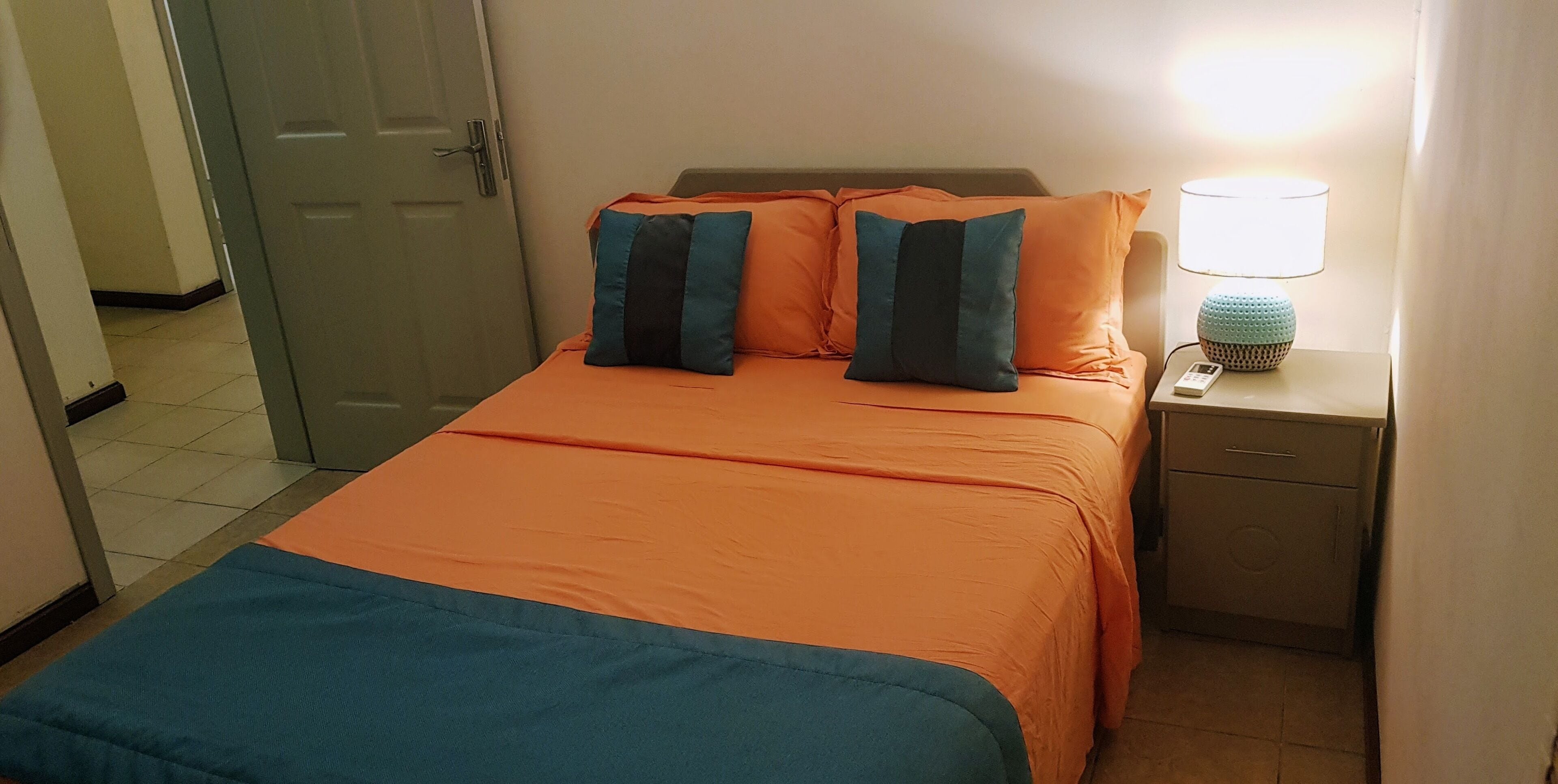 Habitación de hotel con una colcha naranja, dos almohadas, una mesita de noche con una lámpara y una puerta que da a otra habitación.