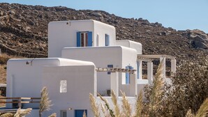 A white building with blue doors and windows, surrounded by dry vegetation and rocky hills. - Electra Village (Mykonos)