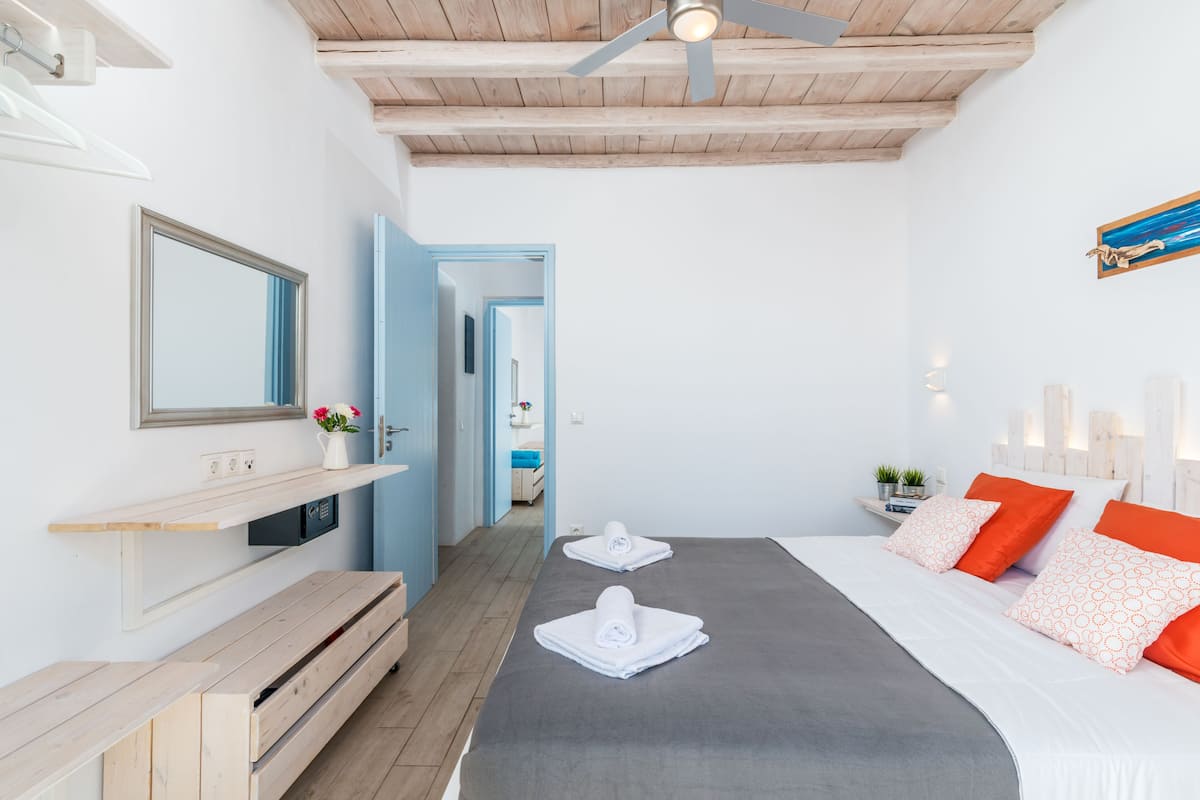 Una habitación de hotel moderna con techo de madera, una cama grande con almohadas anaranjadas y blancas, un banco, un espejo y una pequeña planta en la mesita de noche.