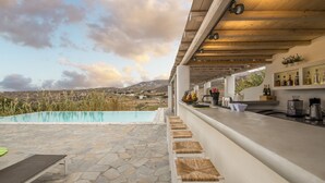 A poolside area with stone flooring, white bar stools, and a bar with shelves of bottles and a coffee machine. - Electra Village (Mykonos)