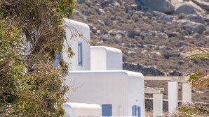 A white building with blue accents, surrounded by olive trees and dry vegetation, set against a rocky hillside. - Electra Village (Mykonos)