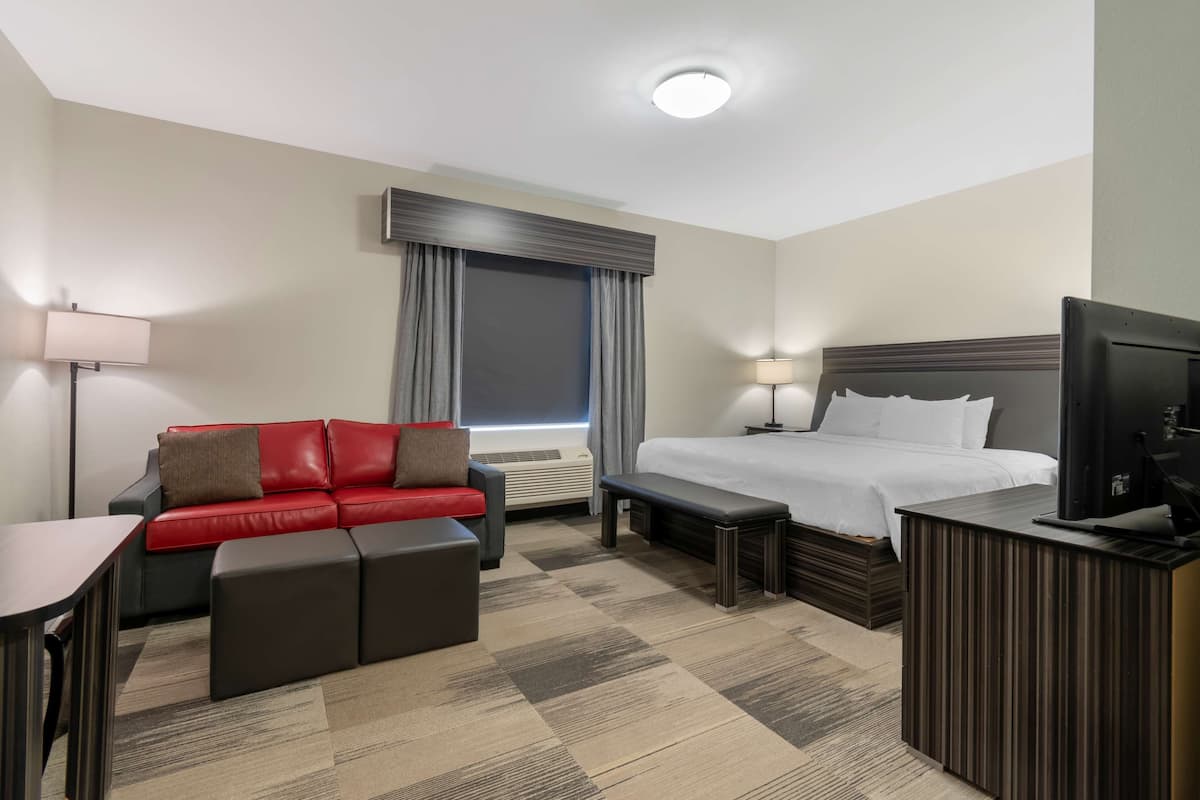 Una habitación de hotel con cama, sofá rojo, escritorio, televisor y ventana con cortinas.