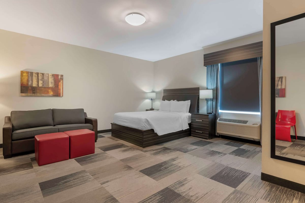 Habitación de hotel con cama, sofá, reposapiés rojo, mesita de noche, lámpara, equipo de aire acondicionado y un cuadro en la pared.