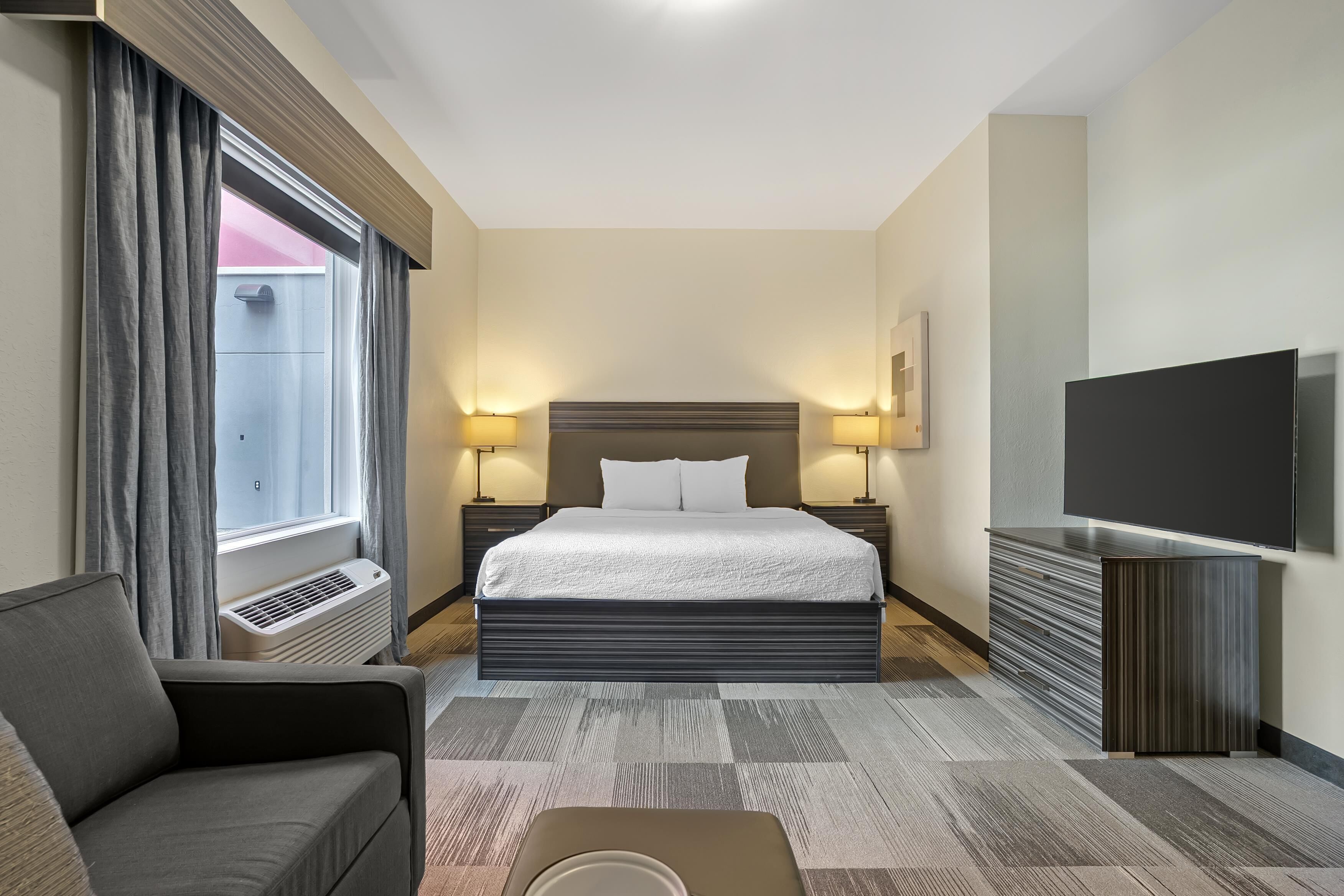 A hotel room with a bed, two bedside lamps, a flat-screen TV, a sofa, and a large window with curtains.