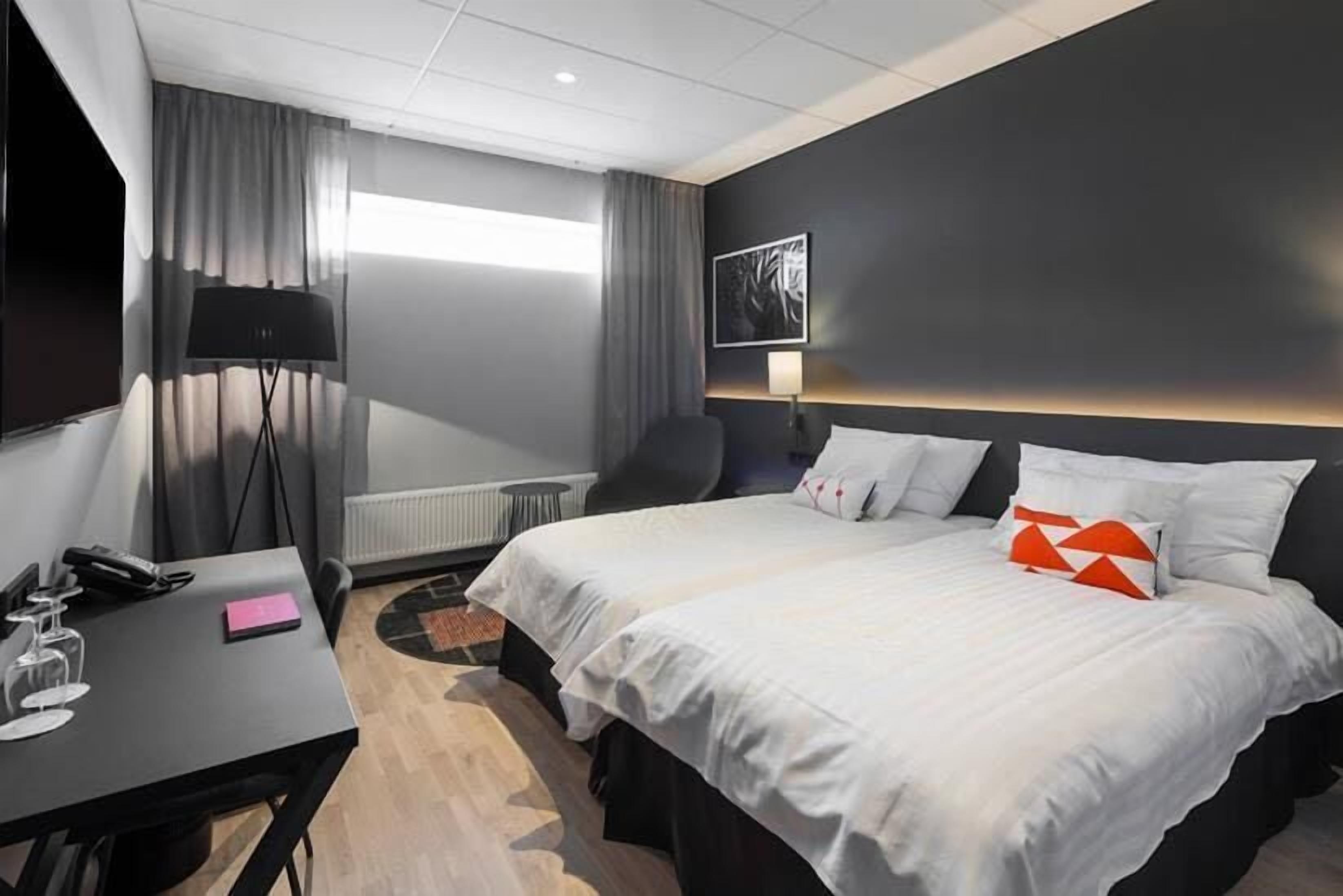Une chambre d’hôtel avec un lit, un bureau, une chaise, une lampe et une fenêtre avec des rideaux.