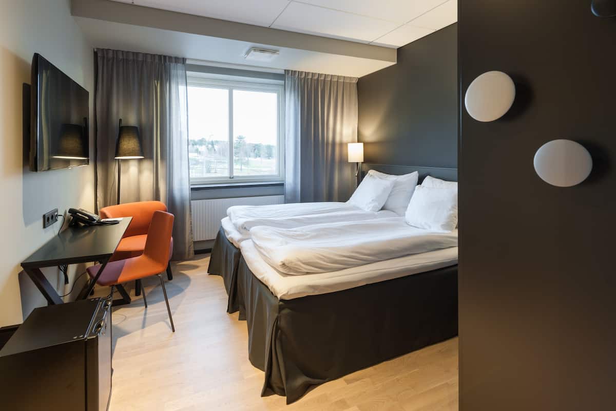 A hotel room with a bed, a desk, an orange chair, and a window with curtains.