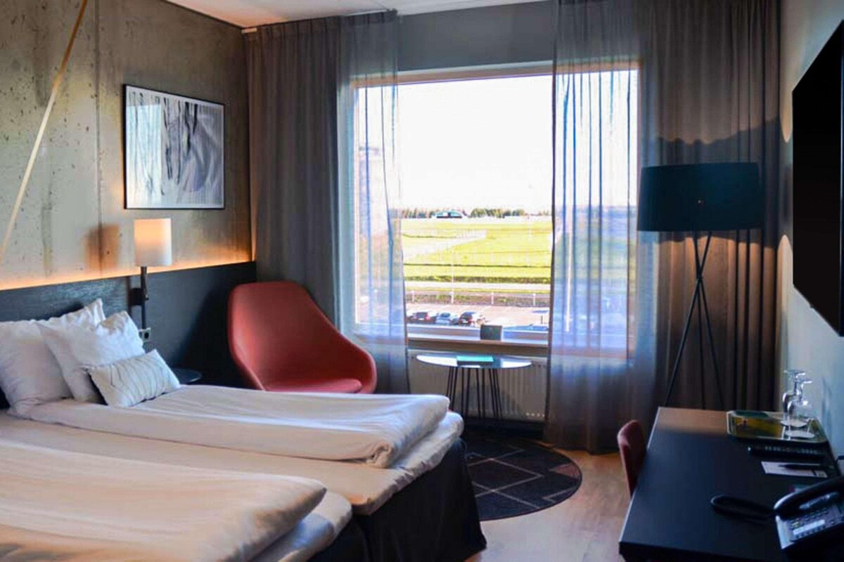 A hotel room with a bed, a red chair, a desk, a lamp, and a view of the outside.