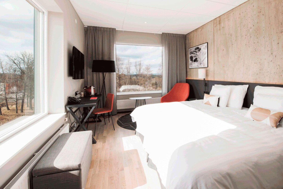A hotel room with a large bed, a desk with a chair, a small table, and a view of the outdoors.