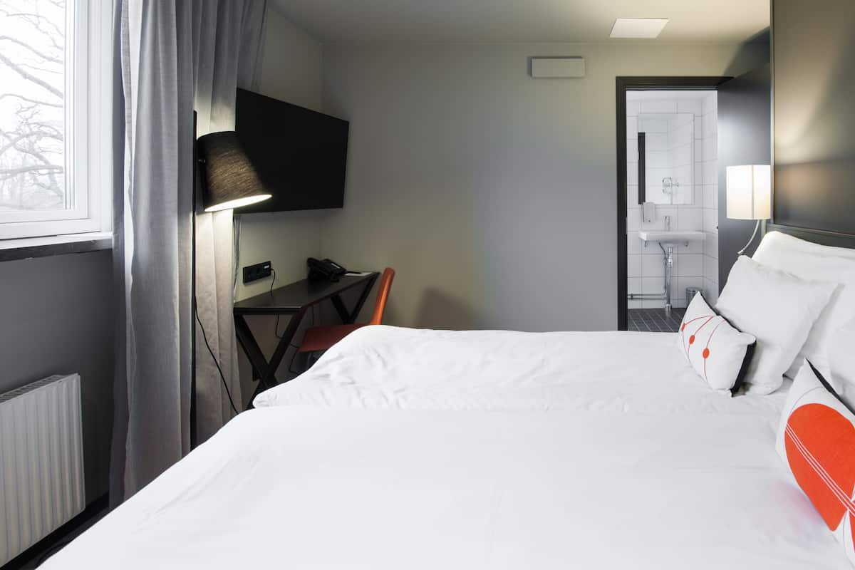 A hotel room with a bed, a desk, a chair, a TV mounted on the wall, and a bathroom visible through an open door.