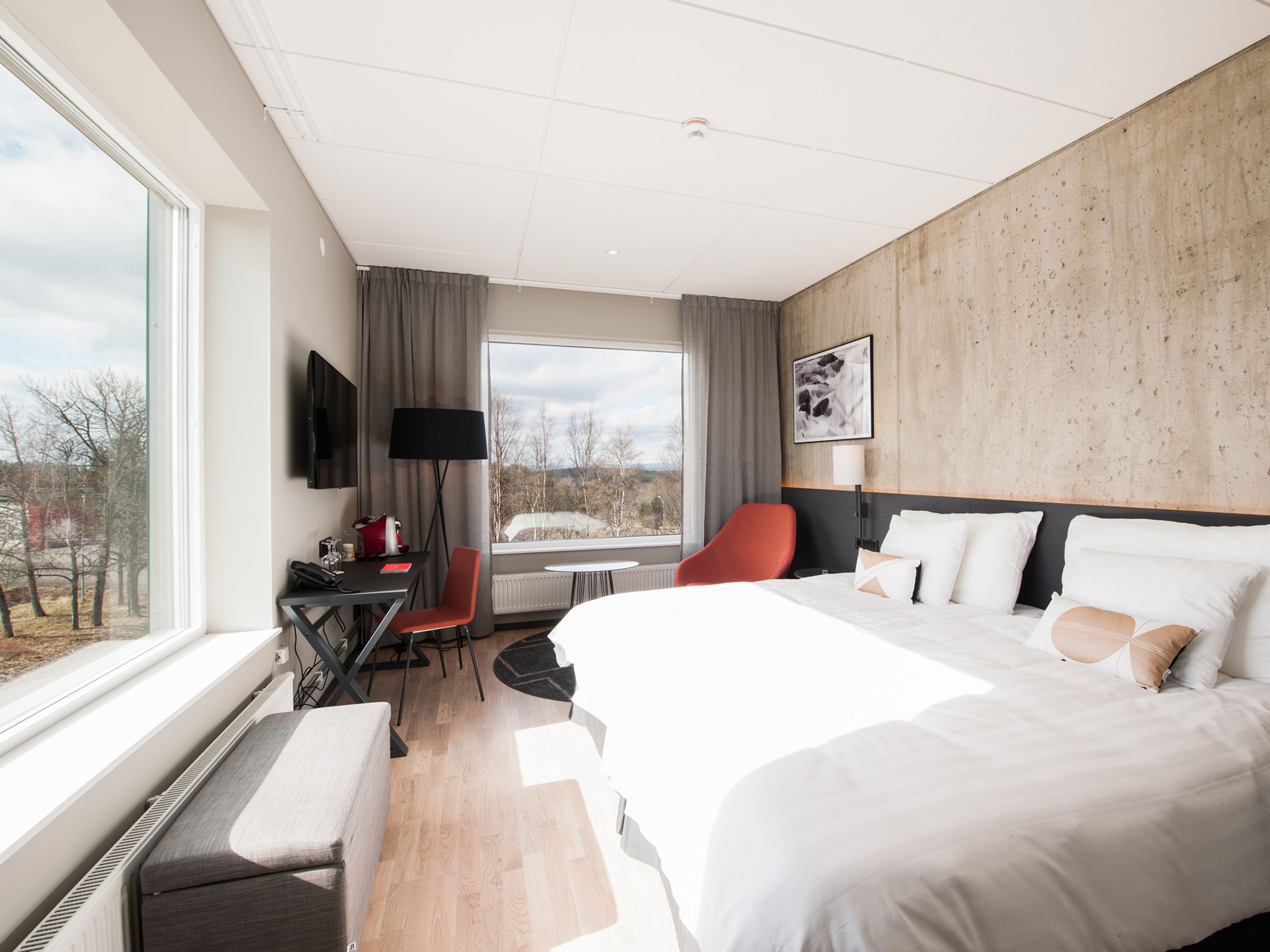 Una habitación de hotel moderna con una cama grande, un escritorio con una silla, un televisor y una ventana con vistas a árboles y edificios.