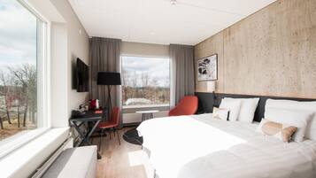 Una habitación de hotel moderna con una cama grande, un escritorio con una silla, un televisor y una ventana con vistas a árboles y edificios.